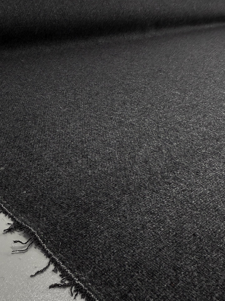 A close-up of Super Cheap Fabrics’ Hopsack Wool - Charcoal - 150cm shows the large wool fabric roll laid flat, highlighting its slightly fuzzy texture and frayed lower left corner.