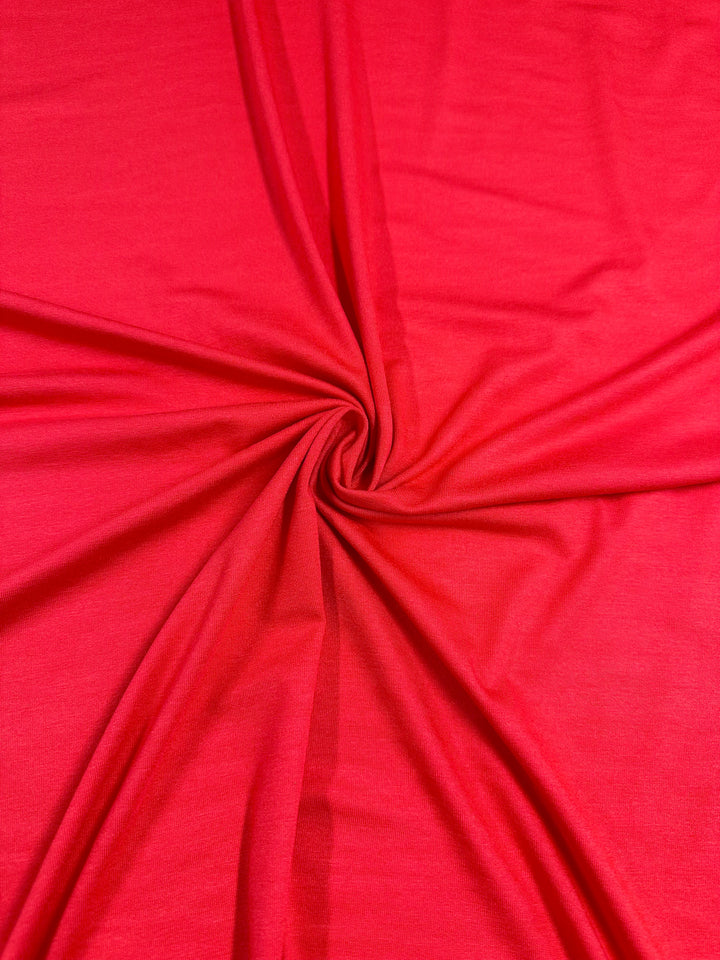 A vibrant red fabric with a soft, smooth texture is gathered and twisted at the center, creating radiating folds and wrinkles that spread outward in all directions. This lightweight two-way stretch fabric in Super Cheap Fabrics' Rayon Lycra - Bittersweet - 145cm adds a touch of shimmer and elegance.