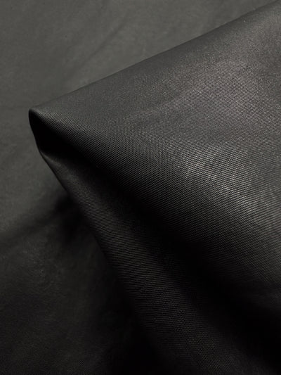 A close-up of Super Cheap Fabrics' Faux Leather - Black - 145cm, showcases its high-end, sleek, and glossy texture with a visible diagonal fold. The slightly reflective finish suggests a synthetic or coated medium-weight material.