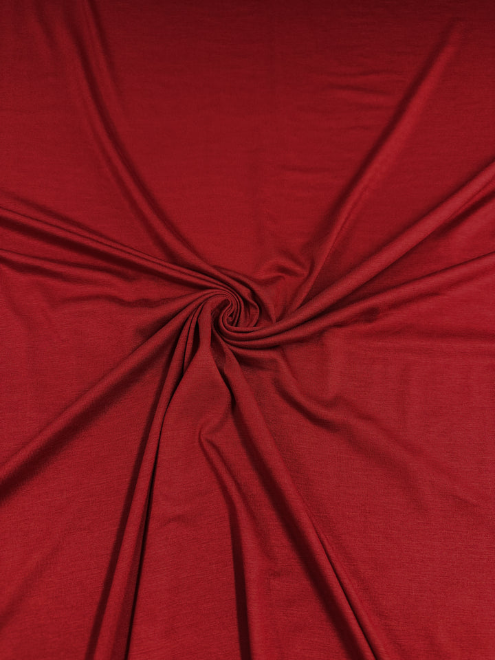 A close-up of deep red Rayon Lycra - Salsa - 145cm fabric with a central twist, creating star-like folds radiating outward. The medium weight fabric appears smooth and slightly reflective, emphasizing its texture and two way stretch. This product is offered by Super Cheap Fabrics.