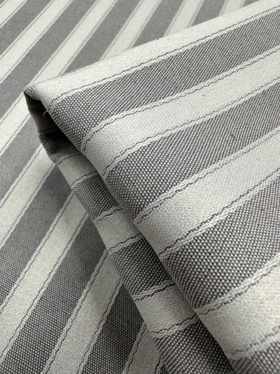 A close-up of folded medium weight fabric from Super Cheap Fabrics shows the Upholstery - Silver & Grey - 140cm. The 100% polyester features alternating light gray and off-white stripes, presenting a textured appearance and neatly showcasing the weave and design.