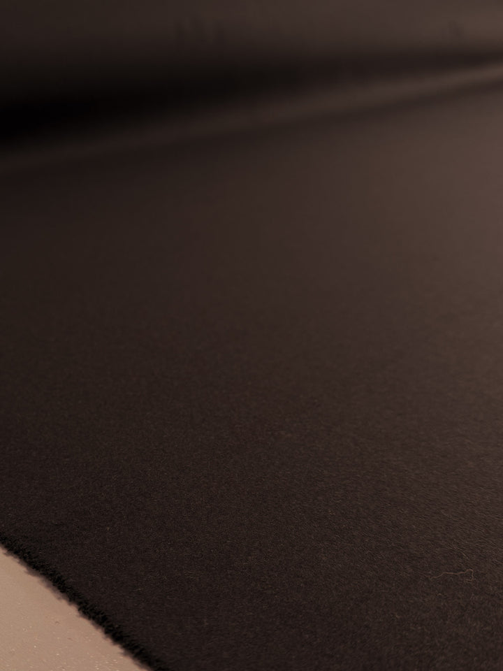 A close-up of Super Cheap Fabrics’ Melton Wool - Black Coffee (150cm) shows its smooth, dark brown texture under soft lighting, with the edge visible in the lower left—a premium outerwear fabric.
