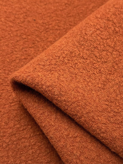 A close-up of the Wool Boucle - Burnt Orange - 135cm fabric by Super Cheap Fabrics reveals its textured, woolly appearance. The fabric is folded at an angle, showcasing its heavy weight and layered thickness which emphasize its cozy feel, making it ideal for overcoats.