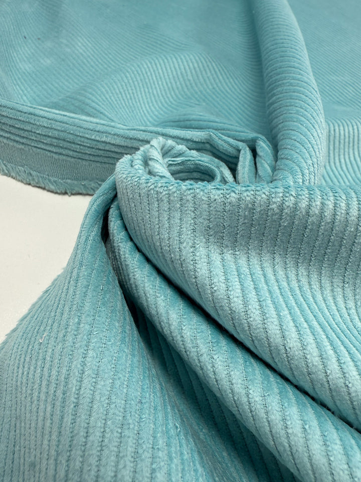 Close-up of Super Cheap Fabrics' Mid Wale Corduroy in pastel turquoise (145cm), showing its signature ribbed texture. The cotton fabric is partially rolled and draped, with soft, velvety ridges and folds highlighted.