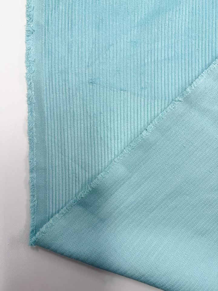 A close-up of Super Cheap Fabrics' Mid Wale Corduroy in Pastel Turquoise (145cm), showing its textured, ribbed surface on one side and smooth reverse, with frayed edges visible on two sides.