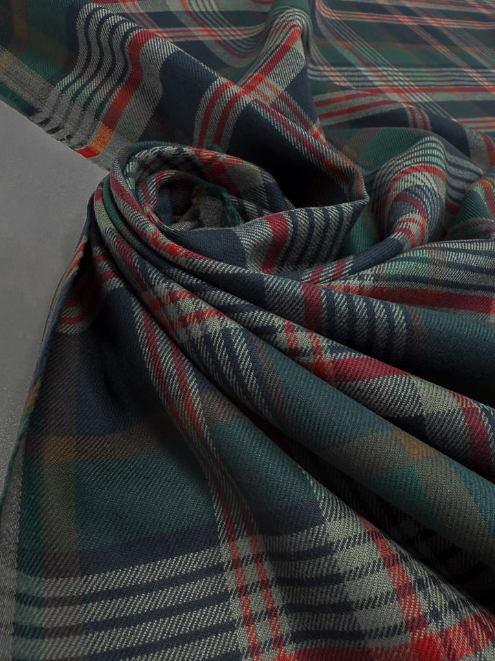 A close-up of Super Cheap Fabrics' Flannel Tartan - Glenmore Forest (145cm) shows soft, plaid cotton flannel in green, blue, red, and beige, draped in gentle folds for a cozy heritage look on a gray surface.
