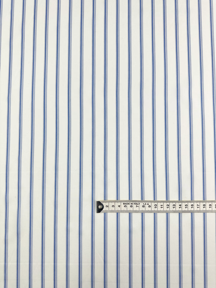 The "GOTS Cotton - Thin Regatta Stripe - 150cm" from Super Cheap Fabrics is a white fabric featuring thin vertical blue stripes that are evenly spaced. Made from certified GOTS organic cotton, the fabric showcases its pattern with a measuring tape placed horizontally along the bottom edge, marked in centimeters and millimeters for precise scale. This fabric is ideal for eco-friendly fashion projects.