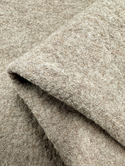 Close-up of a textured, light brown 100% wool fabric, featuring a folded section. The Boiled Wool - Savannah Tan from Super Cheap Fabrics has a heavy weight with a soft and slightly fuzzy appearance, showcasing its cozy and warm nature, perfect for overcoats.