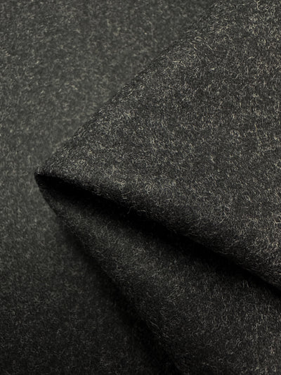 Close-up image of a folded piece of Super Cheap Fabrics' Wool Flannel - Husky - 150cm, showcasing its textured surface and soft, fibrous material. The dark grey fabric appears thick and slightly rough, with fine fibers subtly visible throughout—ideal for making durable coats or jackets.