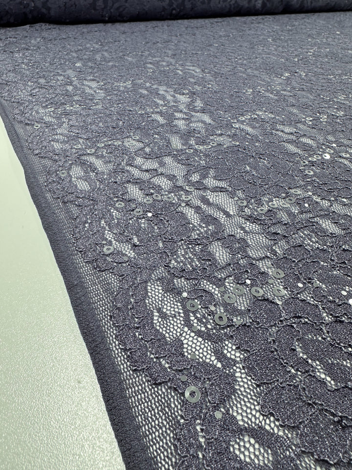 A close-up view of Super Cheap Fabrics' Evening Lace - Queen - 140cm reveals a delicate gray polyester fabric adorned with an intricate floral lace pattern and embedded sequins. The lightweight material is spread out evenly, highlighting its detailed texture and shimmering elements.