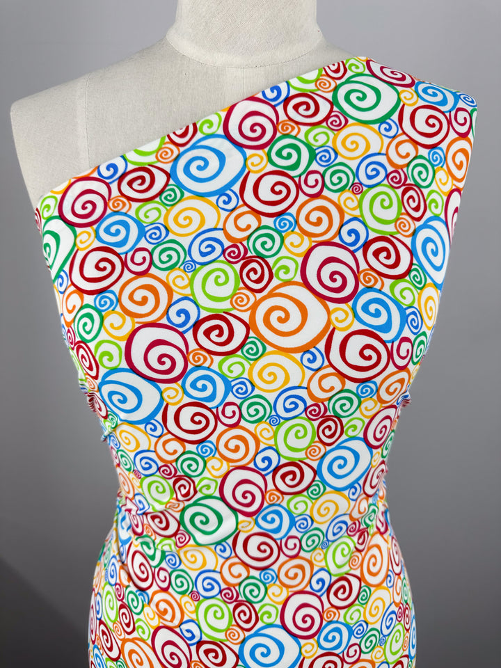 A mannequin is dressed in a vibrant, one-shoulder dress made from Super Cheap Fabrics' Printed Nylon Lycra - Swirly - 155cm, featuring an array of colorful spirals in red, green, blue, yellow, and orange on a white background. The high-quality print on the soft fabric creates a lively and whimsical pattern, adding a cheerful and dynamic look to this multi-use dress.