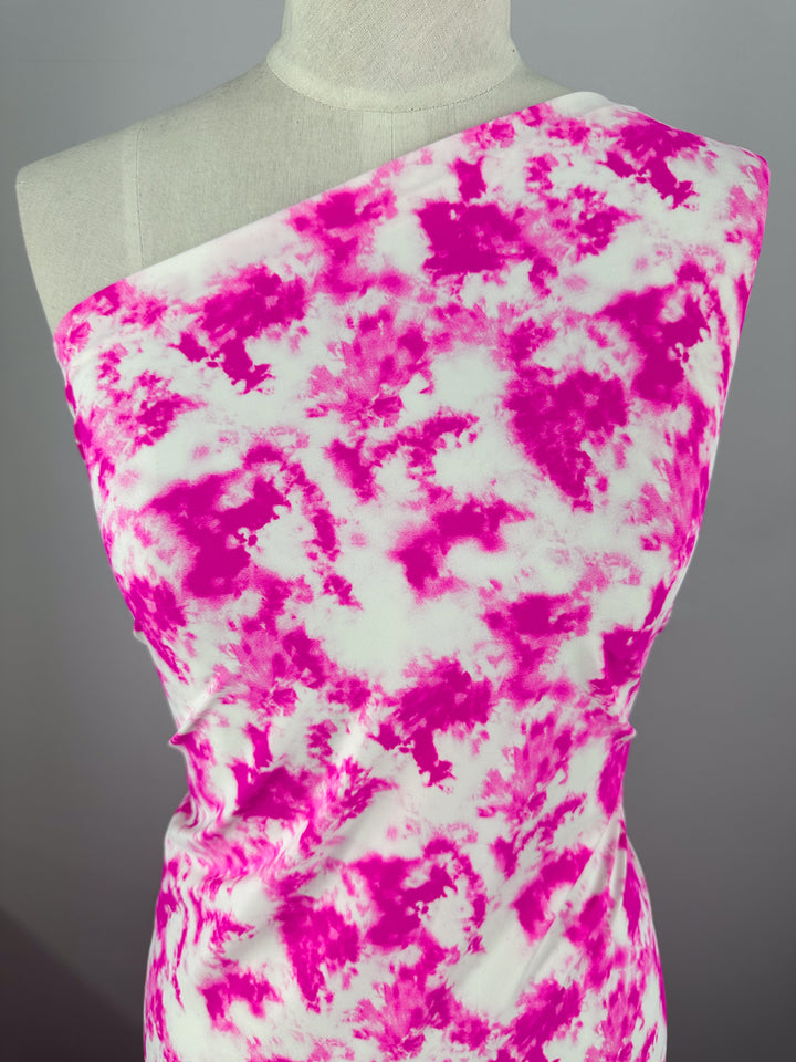 A mannequin is draped with a one-shoulder garment featuring a vibrant pink and white tie-dye pattern. The lightweight fabric, identified as Printed Nylon Lycra - Bandhani - 155cm from Super Cheap Fabrics, has a soft, flowing appearance, showcasing a bold, artistic design. The plain gray background highlights the striking colors and high-quality print of this versatile piece of clothing.