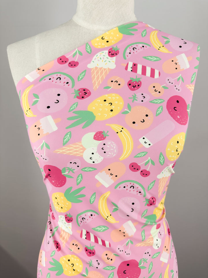 Displayed on a mannequin is a fitted pink dress with a one-shoulder design, crafted from Super Cheap Fabrics' Printed Nylon Lycra - Fruit Salad - 152cm. The soft fabric boasts a playful, high-quality print featuring an array of cheerful fruits—pineapples, strawberries, kiwis, bananas, and watermelons—as well as ice cream cones, all adorned with colorful and whimsical details.