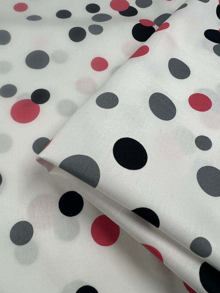 Close-up of Super Cheap Fabrics’ Cotton Sateen - Bordered Bubbles - 145cm, showcasing a 100% cotton fabric with a polka dot pattern featuring black, red, and gray circles of varying sizes on a white background. The lightweight fabric is neatly folded in one corner, making it perfect for household décor items.