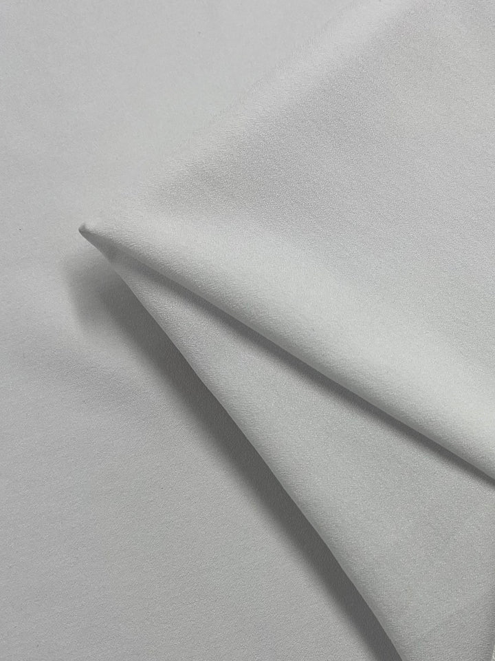 A swatch of Moss Crepe in white, known for its wrinkle-resistant properties, is folded neatly at an angle, showcasing its smooth texture and subtle sheen on a flat grey background. This quality fabric from Super Cheap Fabrics measures 150cm wide.