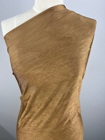 A draped mannequin is covered with a single piece of Shimmer Knit - Bronze - 150cm from Super Cheap Fabrics, resembling the sheen of bronze, creating smooth, soft folds. The background is a plain, light gray color, which contrasts with the warm tone of the fabric.