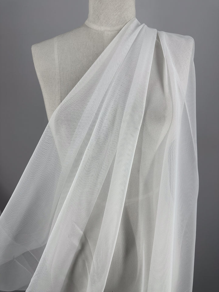 A mannequin displays a draped Soft Tulle - White - 168cm by Super Cheap Fabrics, flowing diagonally across the torso. The plain gray background emphasizes its texture and translucency.