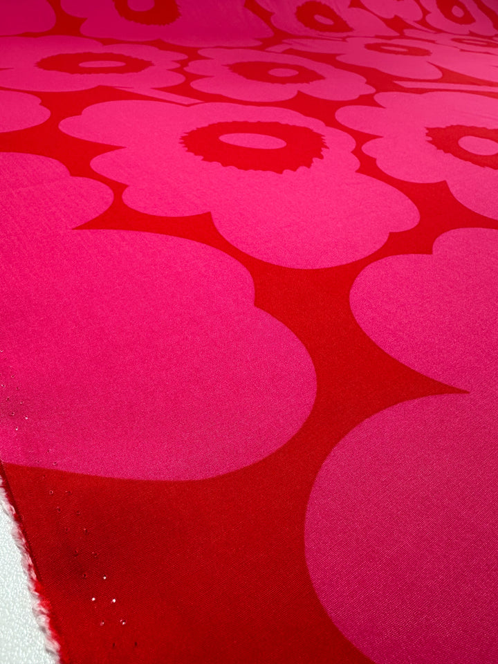 Close-up of Super Cheap Fabrics' Designer Rayon - Jam Poppy (145cm), showing bold pink and red stylized flowers with rounded petals on a vibrant solid background. The fabric edge appears at the lower left, ideal for fashion or home projects.