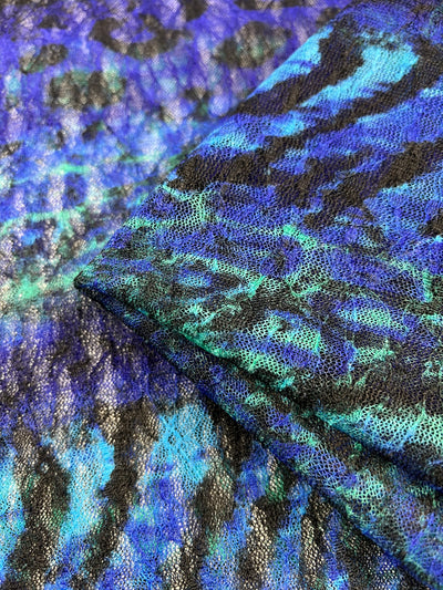 A close-up image of a textured, lightweight sheer fabric with a vibrant pattern of blue, black, and green colors. The Evening Lace - Avatar - 140cm from Super Cheap Fabrics has a slightly glossy appearance, with a folded section on top of a flat one, showing the intricate design prominently—perfect for dresses and skirts.