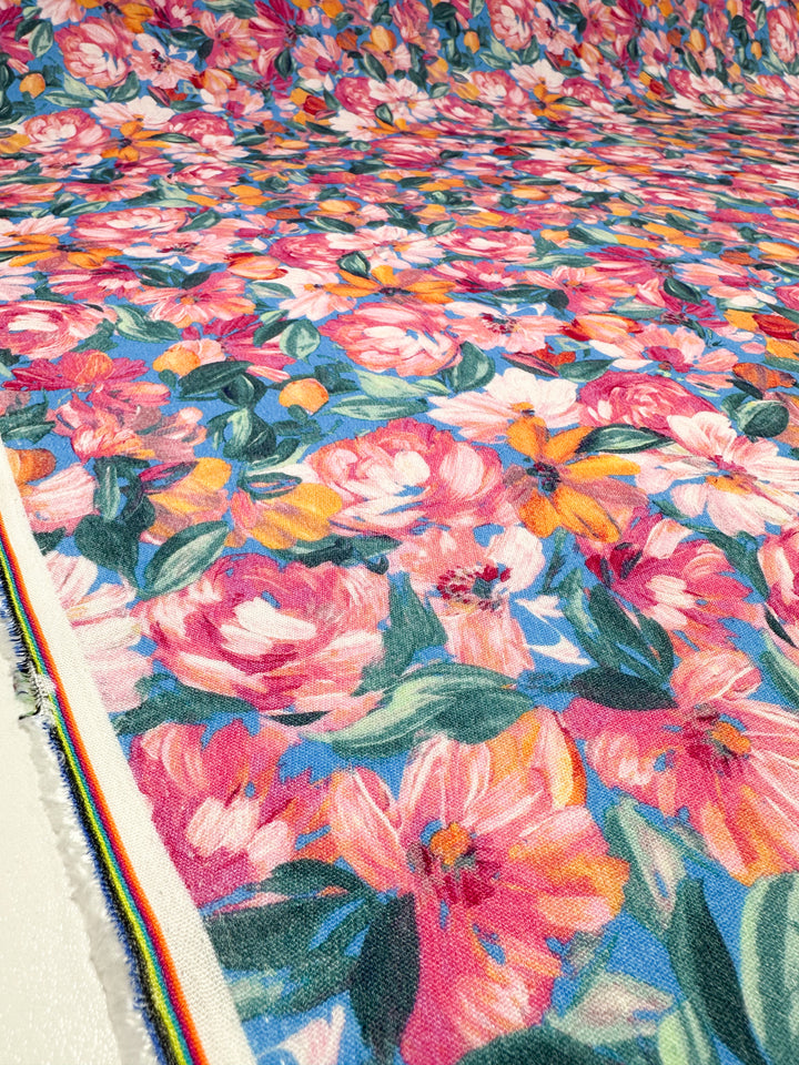 A close-up of Soft Cotton - Acrylic - 140cm from Super Cheap Fabrics displays a stunning floral pattern with large blooms in shades of pink, orange, yellow, and green leaves. The vibrant design extends across the entire breathable fabric, with the edge showcasing a neatly stitched strip.