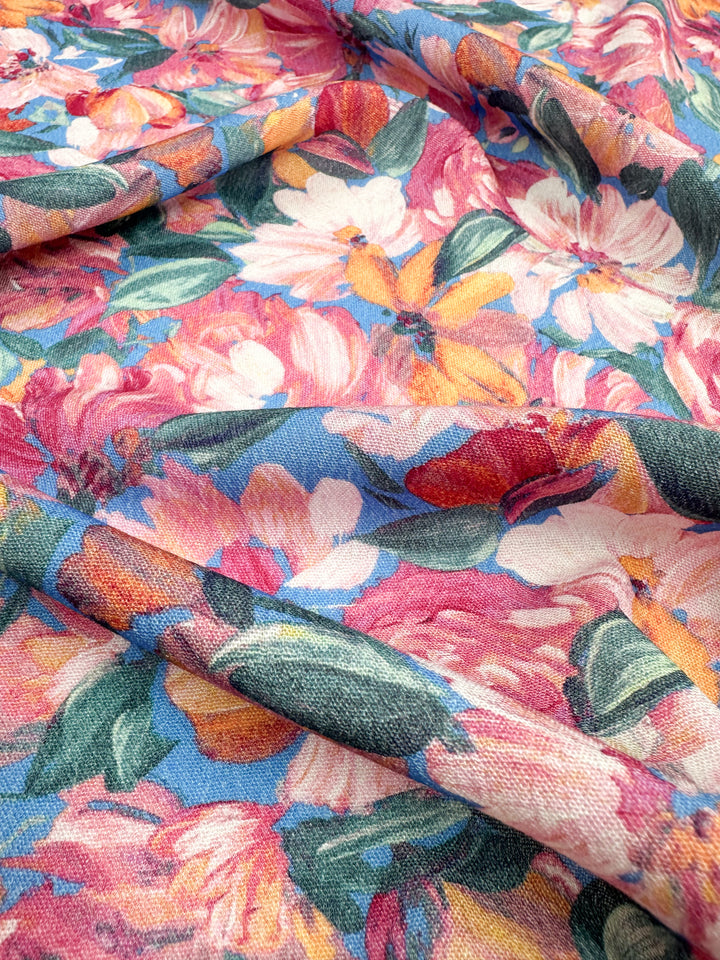 A close-up view of Super Cheap Fabrics' Soft Cotton - Acrylic - 140cm reveals a vibrant, summer fabric. The material, which combines soft cotton and acrylic, features an array of colorful flowers in shades of pink, red, yellow, and white with green leaves on a light blue background. The 100% cotton-blend fabric appears to be textured and slightly wrinkled.