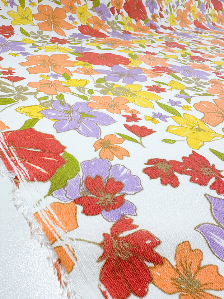 The Soft Cotton - Illustrator - 140cm from Super Cheap Fabrics features a lively floral design on a white background, adorned with an array of flowers in vibrant shades of red, orange, yellow, purple, and green. This summer fabric is made of soft cotton and has a slightly frayed edge, adding texture and character to the material.