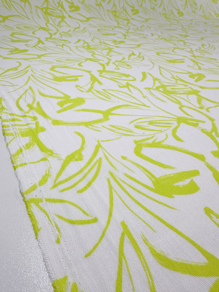 A close-up view of the Soft Cotton - Love Bird - 140cm fabric from Super Cheap Fabrics showcases a white background adorned with bright yellow abstract leaf patterns. Made from soft cotton, the fabric is spread out smoothly, creating a vibrant, dynamic visual effect.