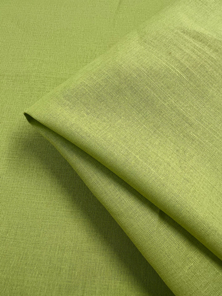 Close-up of a neatly folded piece of Pure Linen - Daiquiri Green - 140cm by Super Cheap Fabrics. The 100% linen material showcases a fine texture with subtle, visible weaves and fibers. The image focuses on the folds and texture, emphasizing the lightweight fabric's softness and quality.