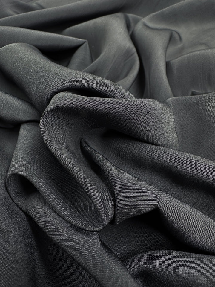 A close-up of the Designer Microfibre - Charcoal, a product by Super Cheap Fabrics, displays its dark, silky polyester arranged in soft, flowing waves to highlight its smooth texture and elegant drape.