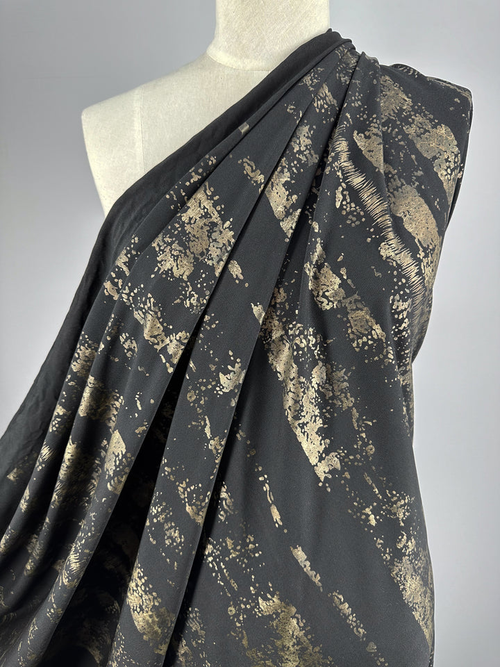 A mannequin adorned in the Evening Lycra fabric by Super Cheap Fabrics, showcasing a medium-weight blend of Polyester and Spandex with a metallic gold abstract splatter resembling brush strokes. The material, known as Rolled Gold, is elegantly draped to cover the top and flows gracefully.