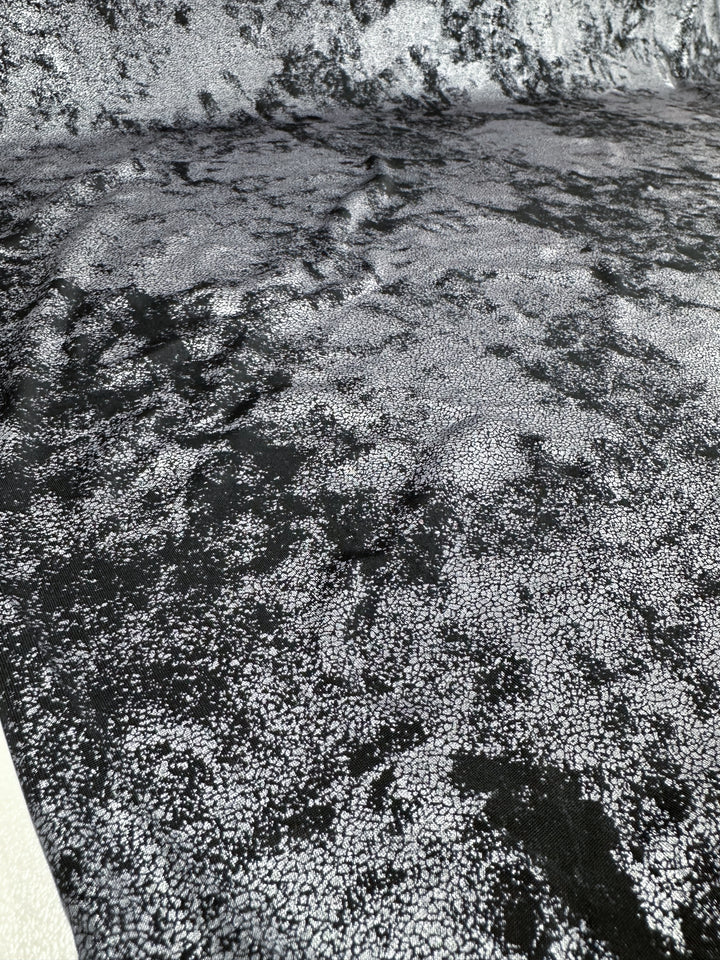 This image showcases a close-up of the medium-weight fabric called "Evening Lycra - Quake - 140cm" by Super Cheap Fabrics. It features a black and white speckled design on a shiny, velvet-like surface, which resembles a mottled or marbled effect. This pattern adds depth and movement to the Polyester/Spandex material.