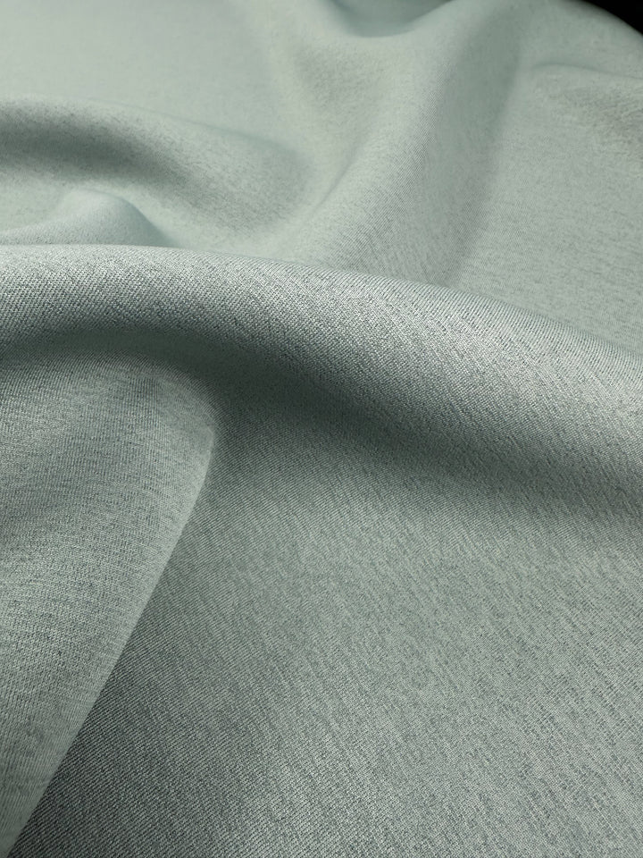 Close-up of the Super Cheap Fabrics' "Scuba Knit - Mermaid - 165cm" in a light gray hue with a slightly heathered texture. The stretchy textile is gently draped, creating soft folds and shadows that convey its softness and flexibility.