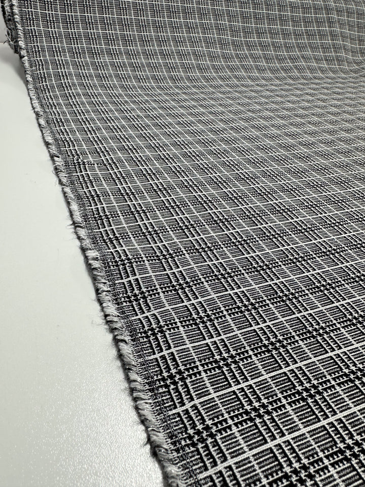 A close-up view of Super Cheap Fabrics' Textured Bengaline - Plaidly - 150cm, a medium-weight fabric showcasing a black-and-white plaid pattern. The multi-use textile is stretched out and angled slightly, revealing a rough, frayed edge on one side. The design features intersecting lines forming a grid pattern.