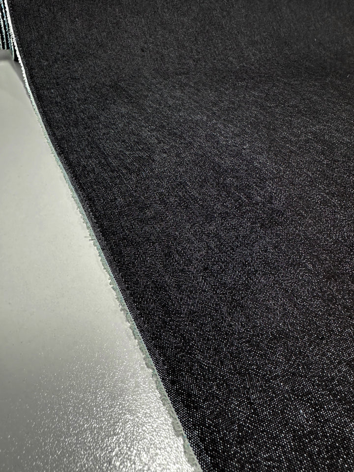 A close-up view of a partially unrolled roll of Lightweight Denim - Black - 135cm from Super Cheap Fabrics, displaying its texture and fine weave. The fabric edge appears frayed, creating a contrast with the smooth, light-colored surface it rests on. The image emphasizes the intricate detail of the 100% cotton material.