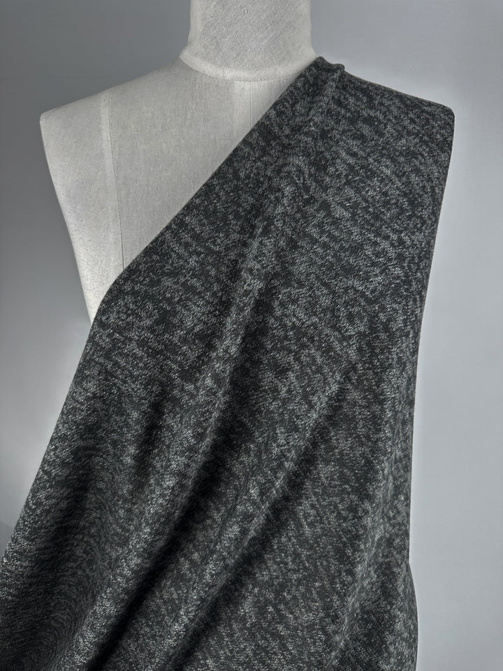 A gray dress form displays Super Cheap Fabrics' Textured Knit - Black Marle - 150cm, draped from one shoulder and falling diagonally across the chest, showing off the dark gray textured stretch fabric against a neutral background.