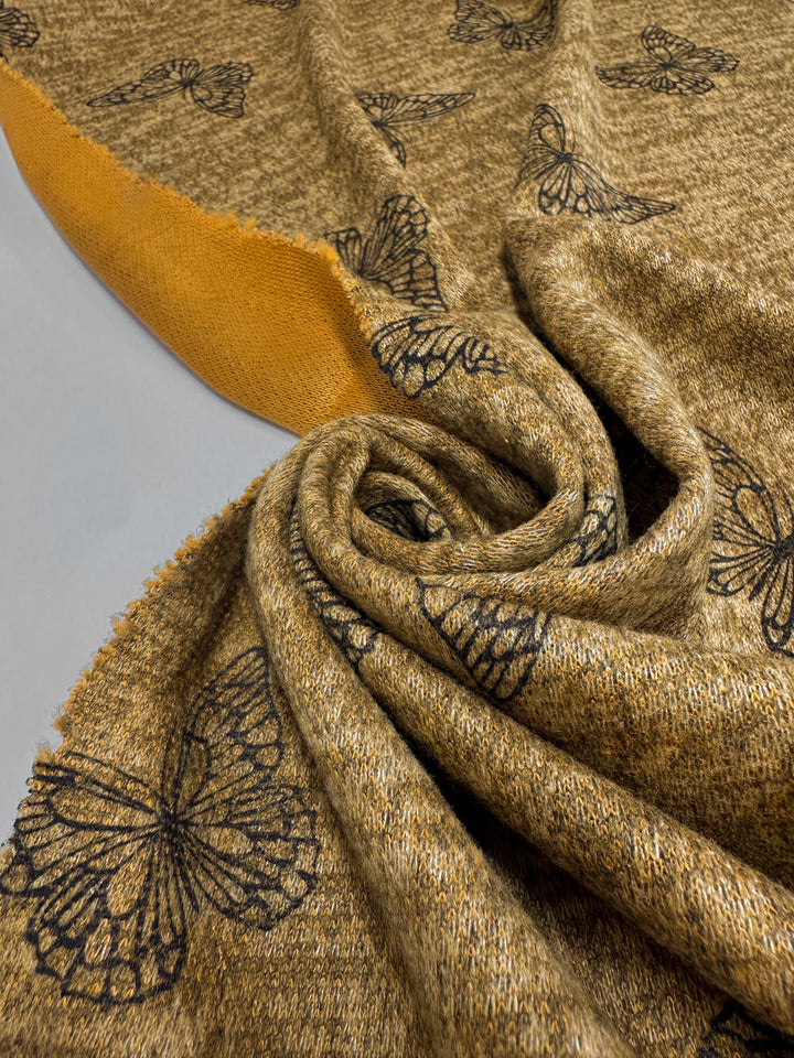 A close-up of Super Cheap Fabrics' Textured Knit - Amber Gold Butterfly - 150cm shows its medium-weight, amber gold fabric with black butterfly prints, swirled to reveal the textured knit surface and plain orange underside.