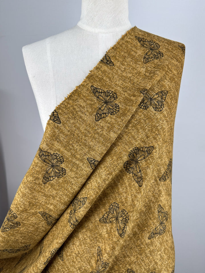 The Super Cheap Fabrics Textured Knit - Amber Gold Butterfly (150cm), a medium-weight fabric with black butterfly outlines, is shown draped over a white dress form against a plain light gray background.