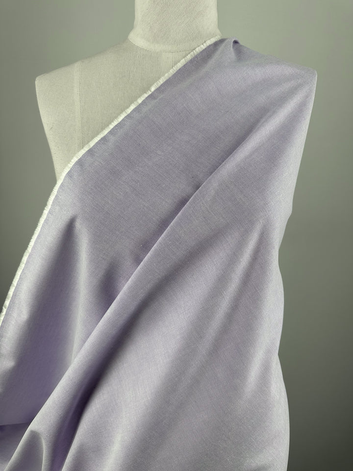 A mannequin draped in Super Cheap Fabrics’ Oxford Cotton - Baby Lavender - 146cm displays its soft, light-to-medium weight fabric and white selvage edge, set against a plain gray background—ideal for versatile garment projects.