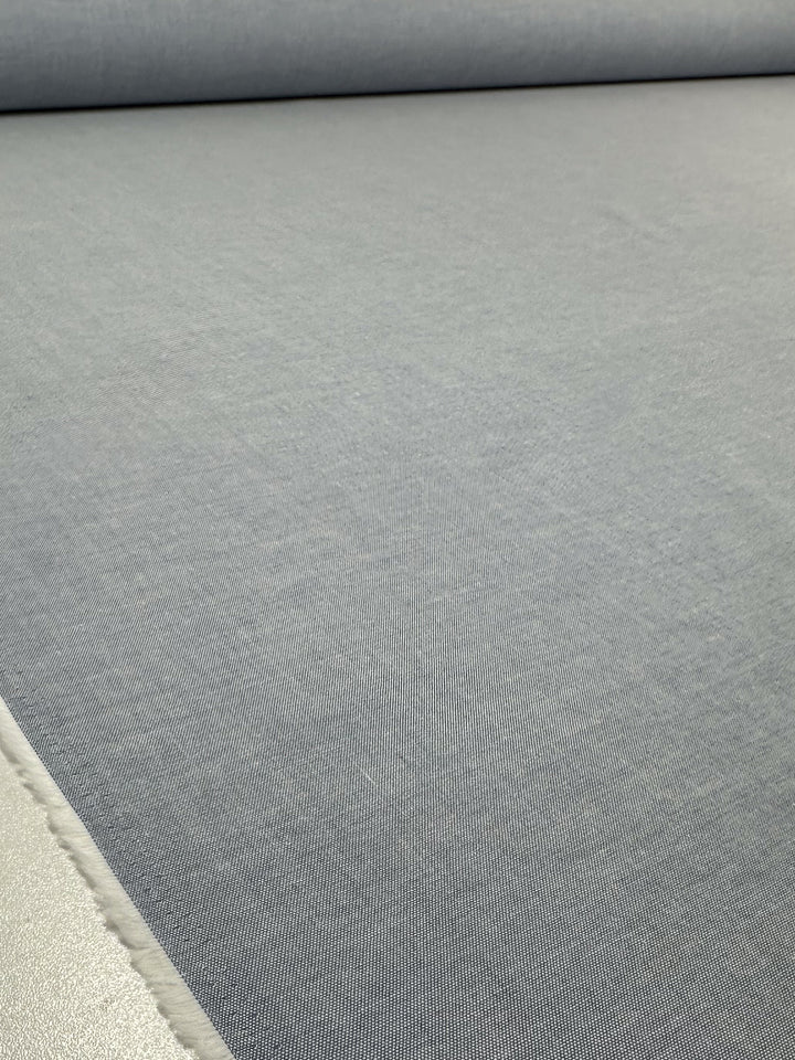 A close-up of Super Cheap Fabrics’ Oxford Cotton - Denim (146cm) showcases its smooth, even light gray texture. Most of the image is filled by the fabric, with an edge visible in the bottom left corner.