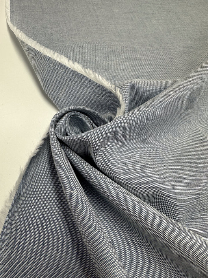 A close-up of Oxford Cotton - Denim (146cm) by Super Cheap Fabrics in light gray, featuring a subtly textured, softly draped fabric with a frayed edge on one side against a white background.