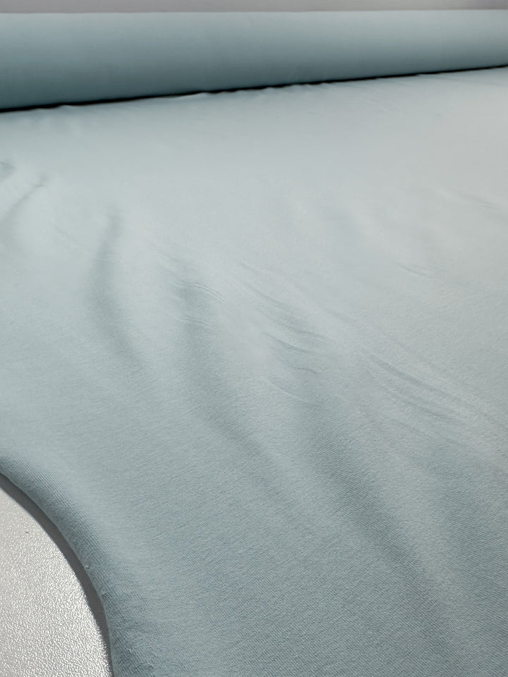 A close-up of Cotton Lycra - Winter Sky - 164cm by Super Cheap Fabrics, partially unrolled to reveal its pale blue color, soft texture, and comfortable feel.