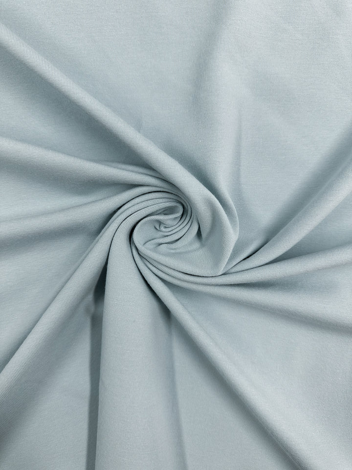 Super Cheap Fabrics’ Cotton Lycra - Winter Sky - 164cm in light gray features a soft texture and gentle stretch. This stylish fabric is shown with swirling folds at the center, highlighting both its comfort and elegant drape.