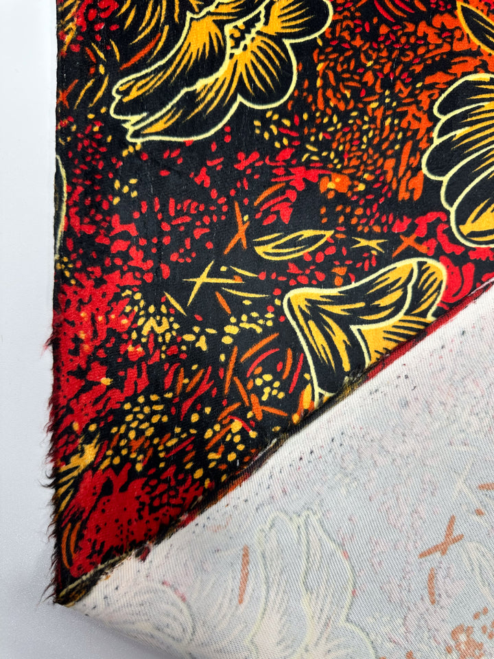 A close-up of DD Velvet - Ember Bloom - 150cm by Super Cheap Fabrics shows its vibrant floral pattern in red, black, yellow, and orange. One corner is folded to reveal the plain white underside with faint printed outlines.