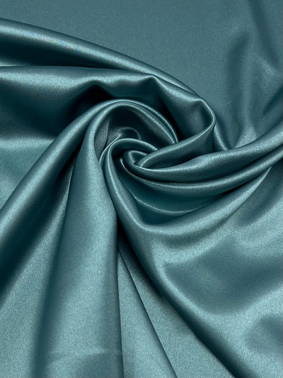A close-up of Super Cheap Fabrics' Satin Deluxe in Nile Blue (150cm), artistically arranged in gentle folds with a centered swirl pattern, showcasing its lustrous and silky texture.
