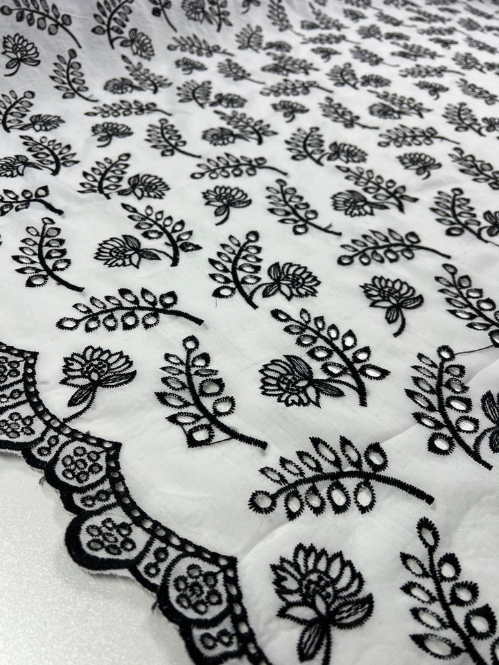 A close-up shot of a white 100% Polyester fabric featuring an intricate black floral and leaf pattern. The fabric has scalloped edges with a detailed lace design, showcasing delicate craftsmanship in the embroidery. The Super Cheap Fabrics Broderie Anglaise - Lotus Leaves - 122cm pattern is repetitive and symmetrical across the material.