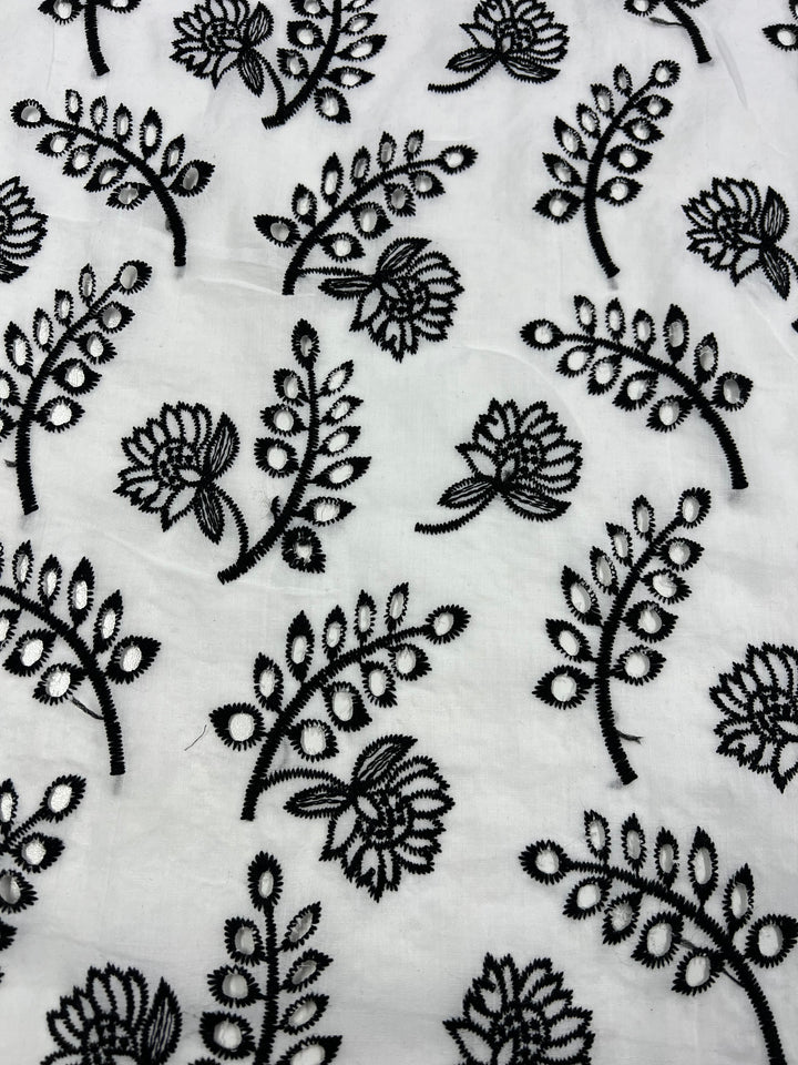 A white fabric featuring a repetitive pattern of black floral embroidery. The design includes flowers with multiple petals and leafy branches, showcasing intricate artistry. This lightweight Broderie Anglaise - Lotus Leaves - 122cm by Super Cheap Fabrics is finely detailed and made from 100% Polyester, creating a visually appealing motif.