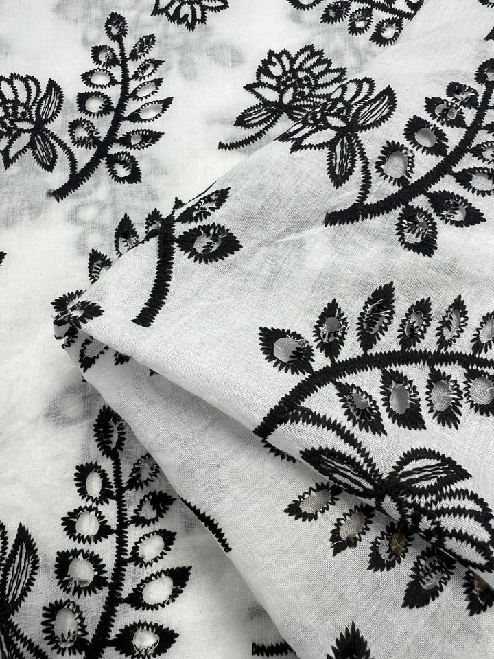 Close-up of Super Cheap Fabrics Broderie Anglaise - Lotus Leaves - 122cm featuring an intricate black floral embroidery pattern with large leaves and buds. The decorative flowers and leaves are sewn with precise and bold stitching, showcasing intricate artistry and adding texture to the soft fabric.