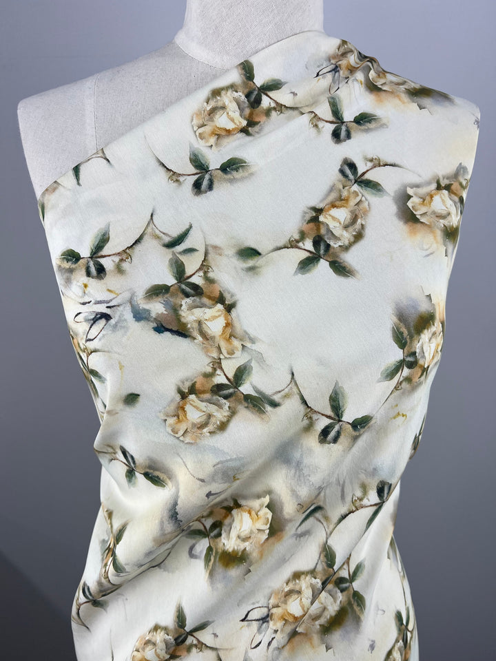 A close-up view of a lightweight fabric draped over a mannequin. The fabric features a multi-colour floral design with beige and white flowers and green leaves against a light, possibly silk, background. The flowers and leaves have a painted, watercolor effect, making it perfect for children's clothing. This is the Designer Cotton - Painted Roses - 145cm by Super Cheap Fabrics.