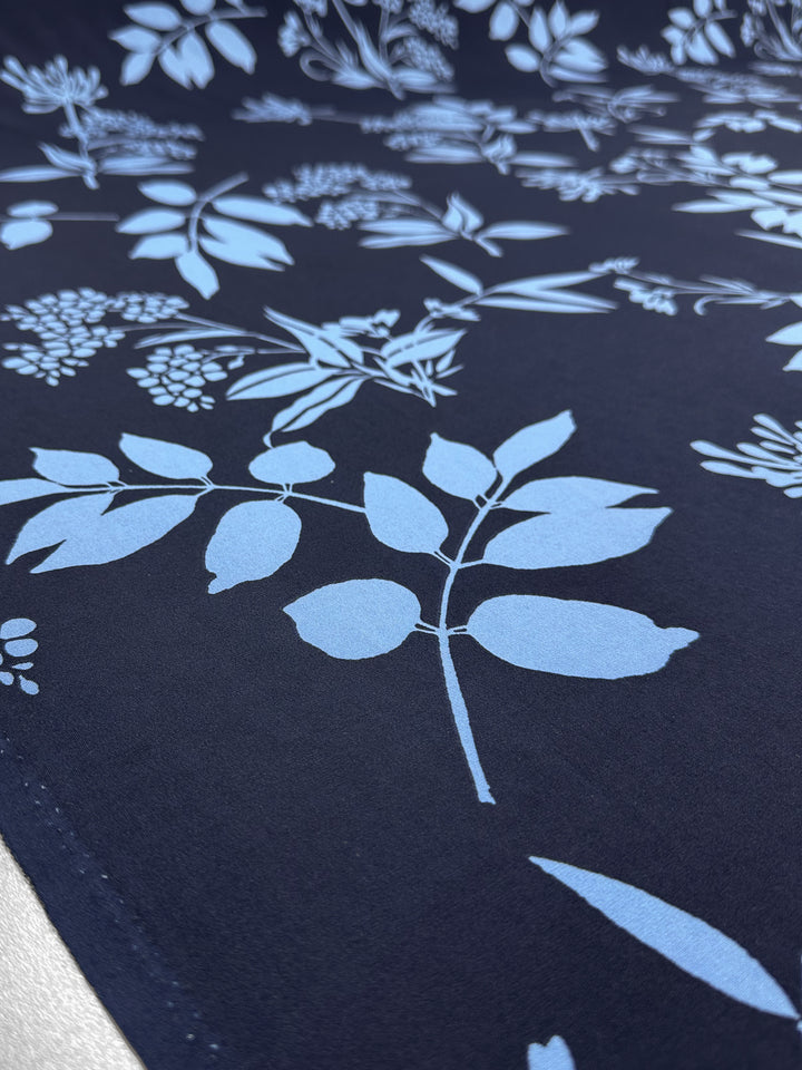 A close-up of Super Cheap Fabrics' Printed Lycra - Silhouette Garden (150cm) shows its dark blue medium-weight lycra with a light blue botanical leaf and flower design, highlighting the fabric's texture and detailed pattern.