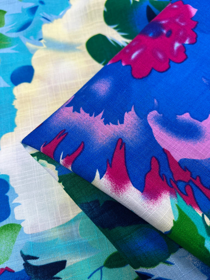 Close-up of two overlapping pieces of Designer Bamboo Rayon - Lantana - 147cm by Super Cheap Fabrics. The top fabric has an intense blue background with vibrant pink, red, and white flowers, while the bottom features a lighter, pastel blue and green floral pattern. Both fabrics have a smooth texture, perfect for versatile clothing with vibrant prints.